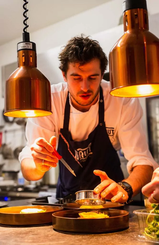 Chef joven concentrado en emplatado gourmet, utilizando una pipeta para aplicar detalles precisos en la cocina de un restaurante, iluminado por lámparas de calor