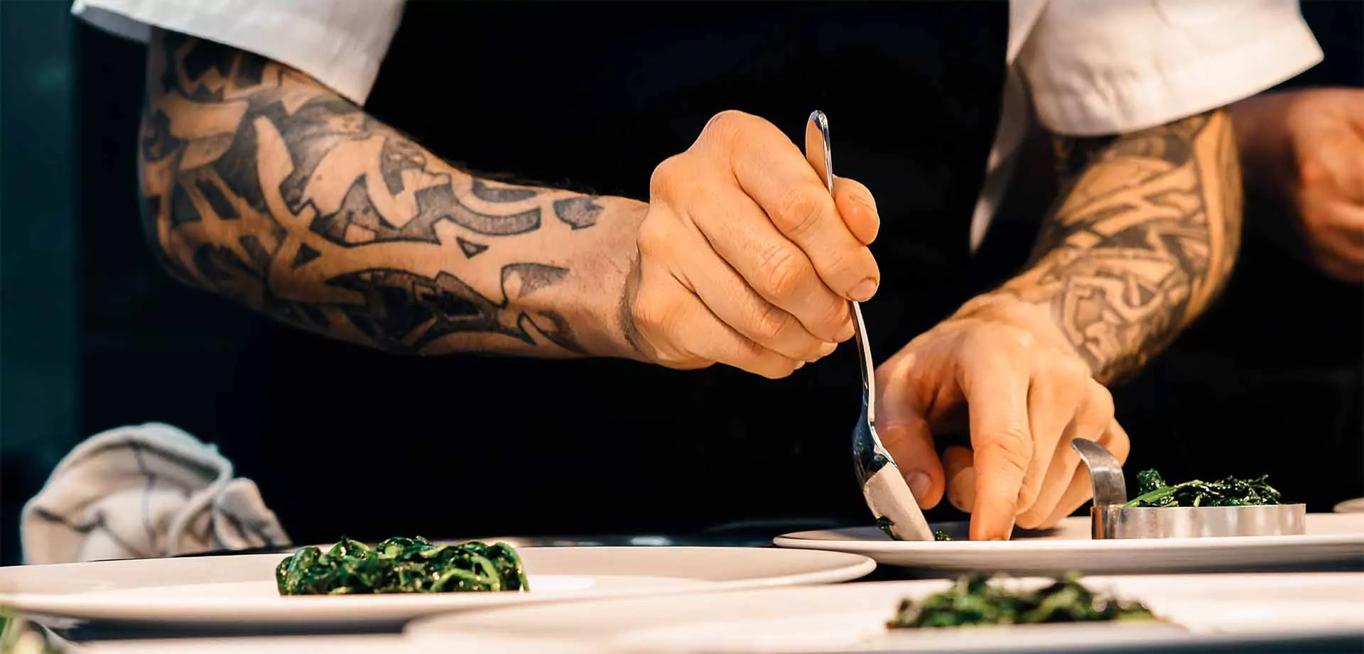 Chef con brazos tatuados emplatando con precisión un plato gourmet en la cocina de un restaurante, utilizando una cuchara y un molde de metal