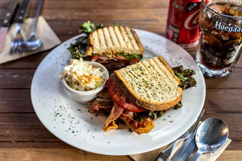 Sándwich BLT con pan tostado, bacon crujiente, tomate y lechuga, servido con ensalada de col y acompañado de refresco en una mesa de madera