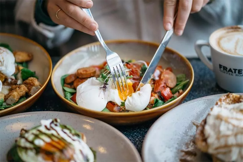 Plato de ensalada con espinacas, chorizo y huevo pochado, con una persona cortando el huevo y café con leche al fondo en un desayuno saludable