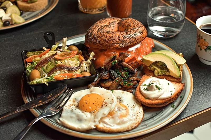Desayuno gourmet con bagel de salmón ahumado, huevos fritos, tostadas con aguacate y queso crema, ensalada fresca con anchoas y champiñones salteados