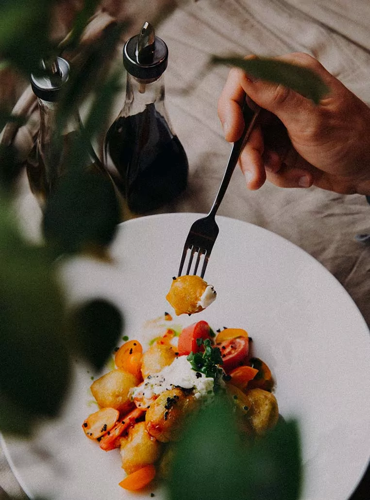 Plato de ñoquis dorados con tomates cherry, queso crema y hierbas frescas, con una mano sosteniendo un tenedor y botellas de vinagre balsámico al fondo Plato de ñoquis dorados con tomates cherry, queso crema y hierbas frescas, con una mano sosteniendo un tenedor y botellas de vinagre balsámico al fondo