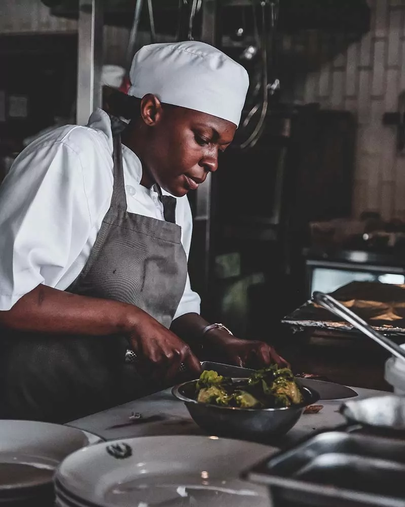 Chef profesional con gorro y delantal preparando una ensalada en la cocina de un restaurante, con utensilios y platos listos para el servicio