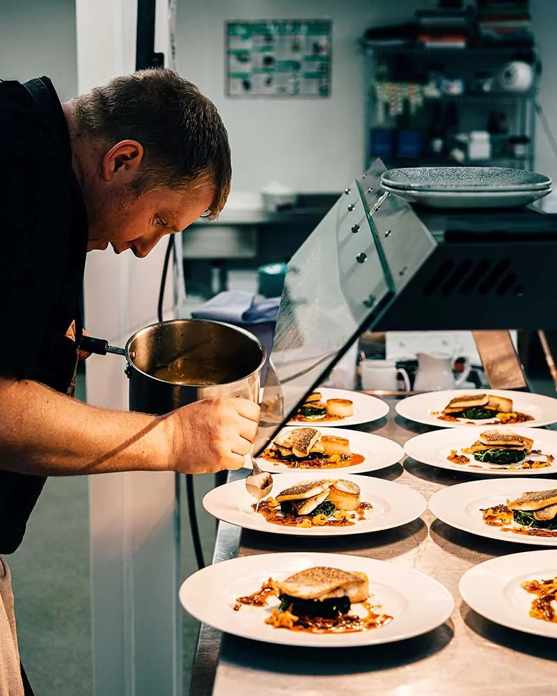 Chef emplatando con precisión una serie de platos gourmet con pescado, papas y salsa en la cocina de un restaurante profesional